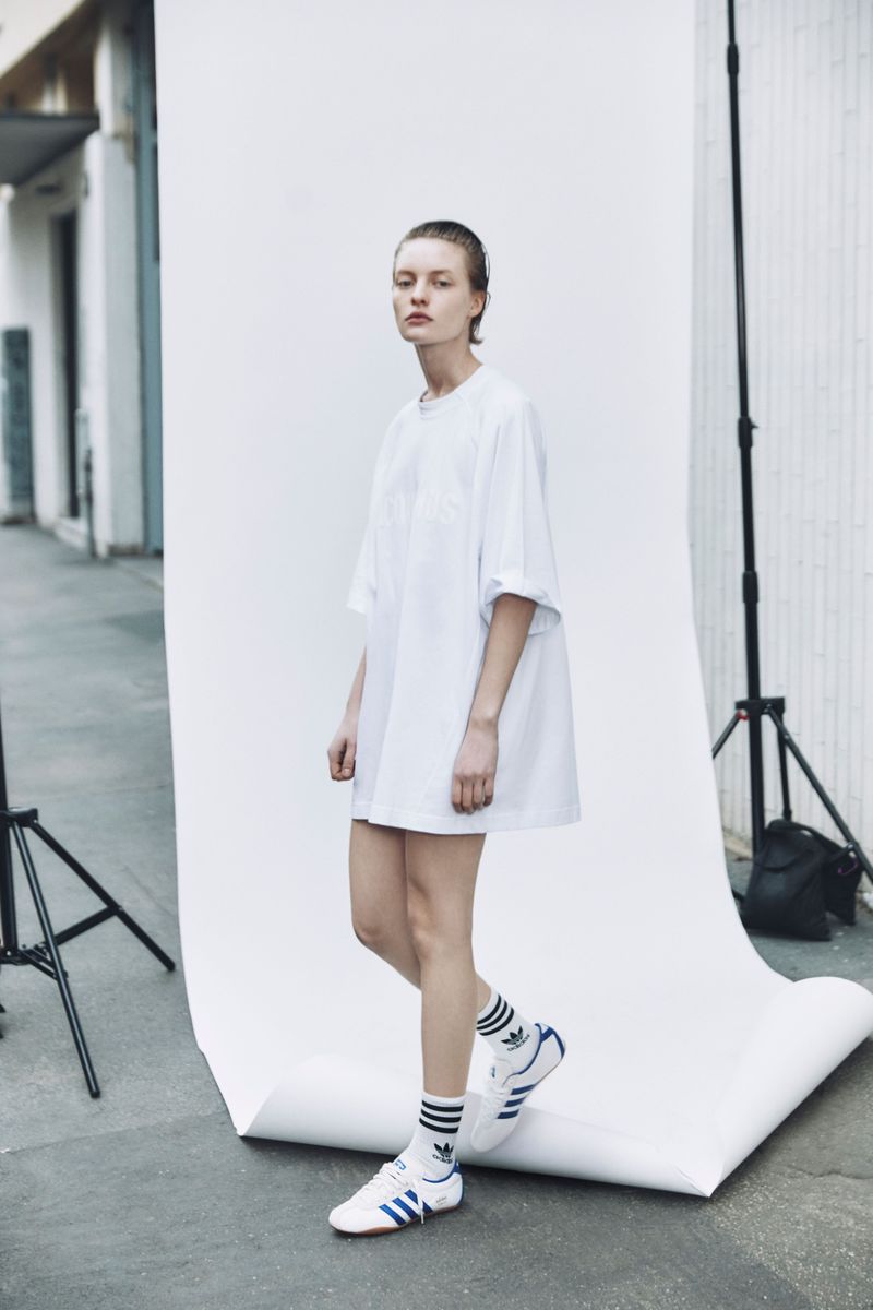 Fashion shot of a slender adult female in a white tee shirt and sneakers, striking a pose in a studio-like setting with a backdrop.
