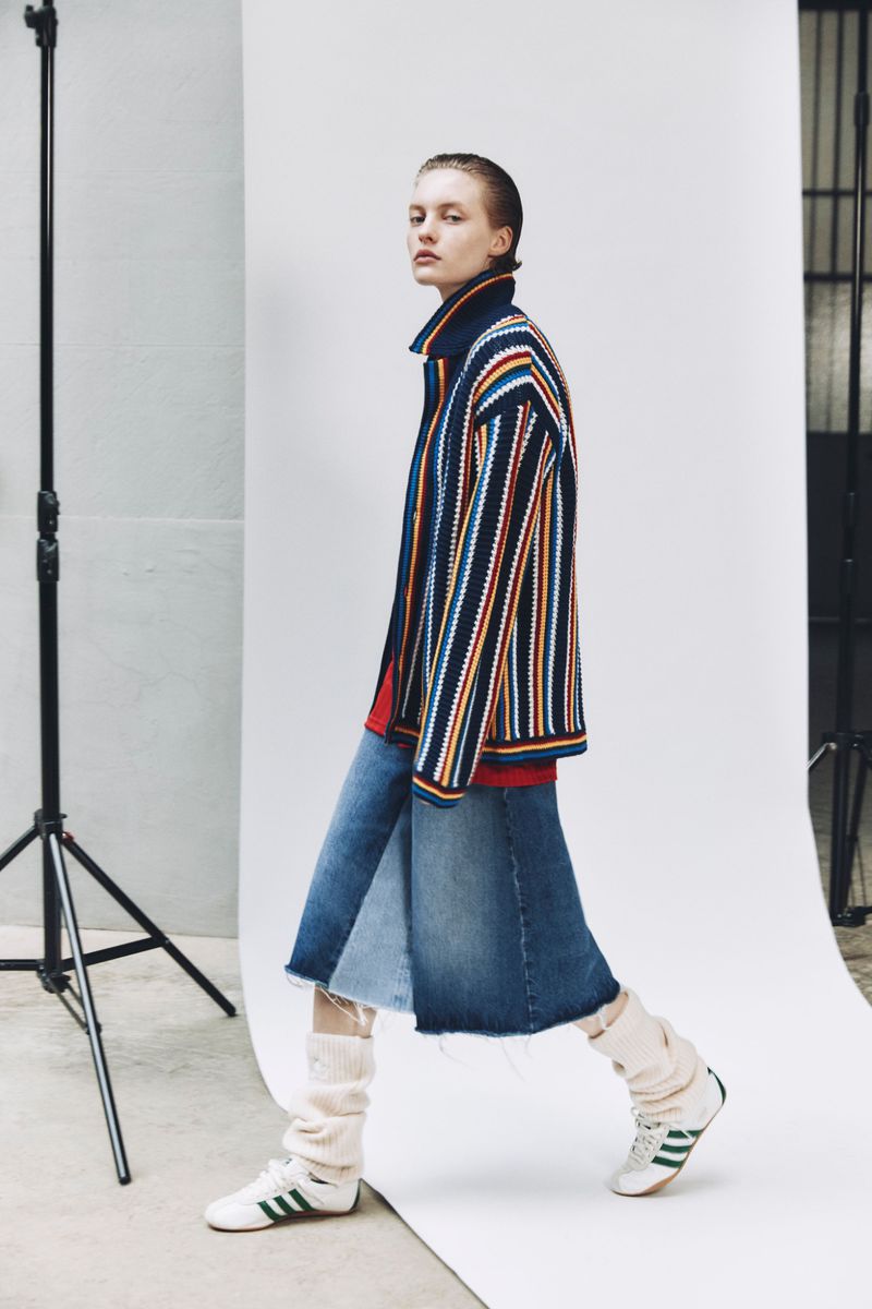 A young woman strides in a fashion shot, wearing a striped jacket, denim skirt, leg warmers, and sneakers, set against a neutral backdrop.