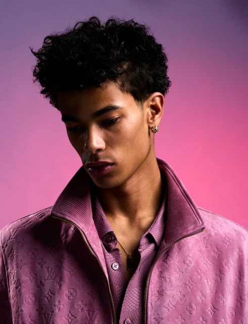 Portrait of a young adult male with curly hair wearing a pink patterned jacket and shirt against a pink and purple gradient background.
