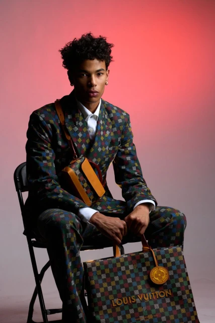 Young adult male in patterned suit sitting with branded tote bag in a studio, posing with serious facial expression against red backdrop.