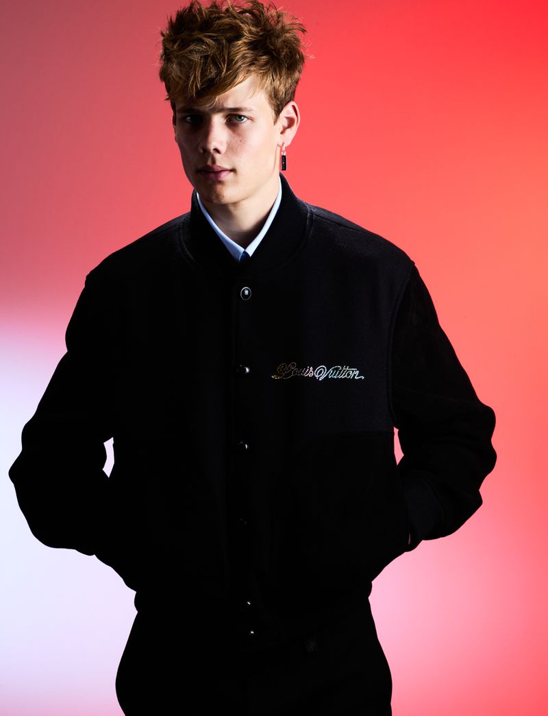 Portrait of a young male model with red hair wearing a black jacket with embroidery against a blended red and white backdrop.