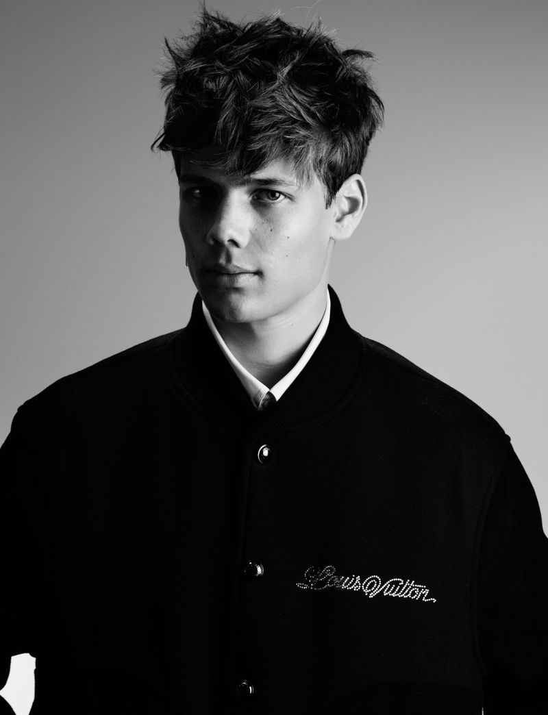 Portrait of a young male model with dark messy hair wearing a black bomber jacket against a plain grey background