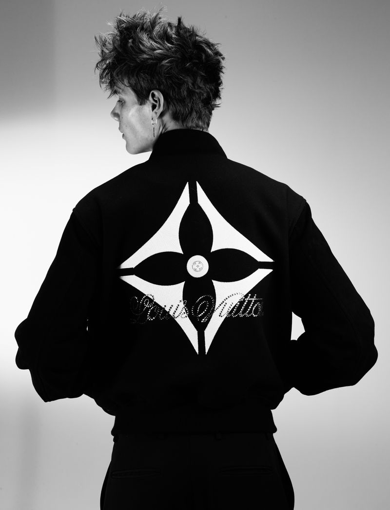 Model seen from the back wearing a black jacket with a large white Louis Vuitton flower logo on the back against a seamless grey backdrop