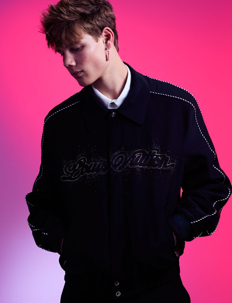 Young adult poses in a navy jacket with decorative white stitching and the 'Louis Vuitton' logo in front of a pink and purple background.