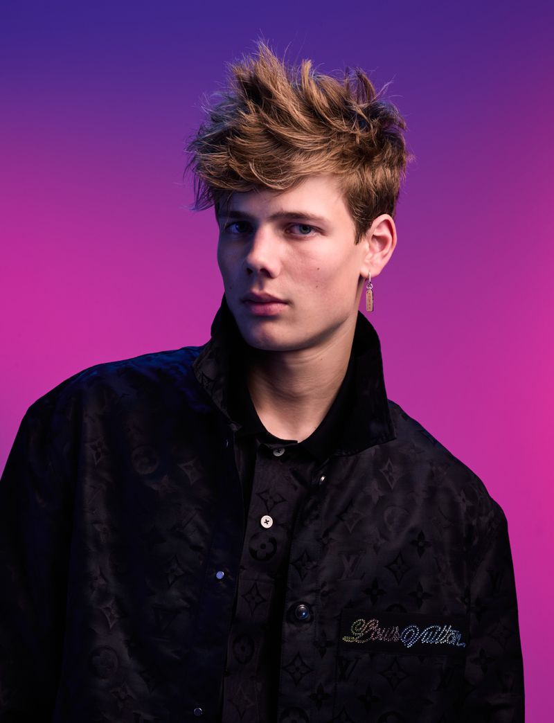 Young male model with tousled brown hair wearing a dark patterned jacket against a vibrant blue and pink gradient background