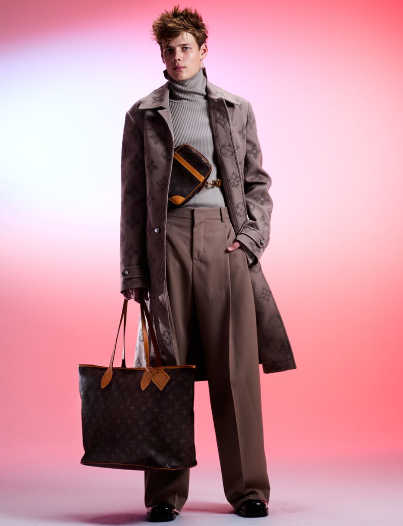 Adult male fashion model poses indoors wearing patterned coat, grey turtleneck, and wide-legged pants, holding monogrammed designer bag.