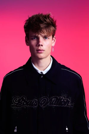 A young adult male with brown hair poses indoors against a pink and purple backdrop, wearing a stylish black jacket with text design.