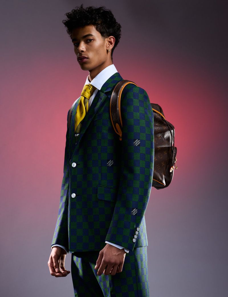 Young adult man poses in a green and blue checked suit with a brown backpack against a pink-toned backdrop in a portrait-style studio shot.