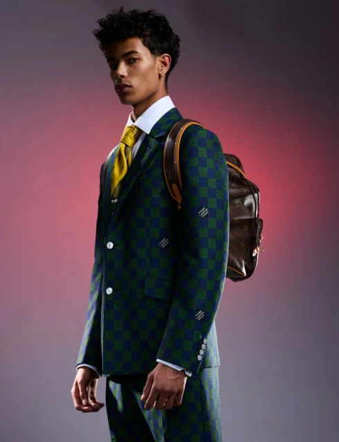 Young adult man poses in a green and blue checked suit with a brown backpack against a pink-toned backdrop in a portrait-style studio shot.
