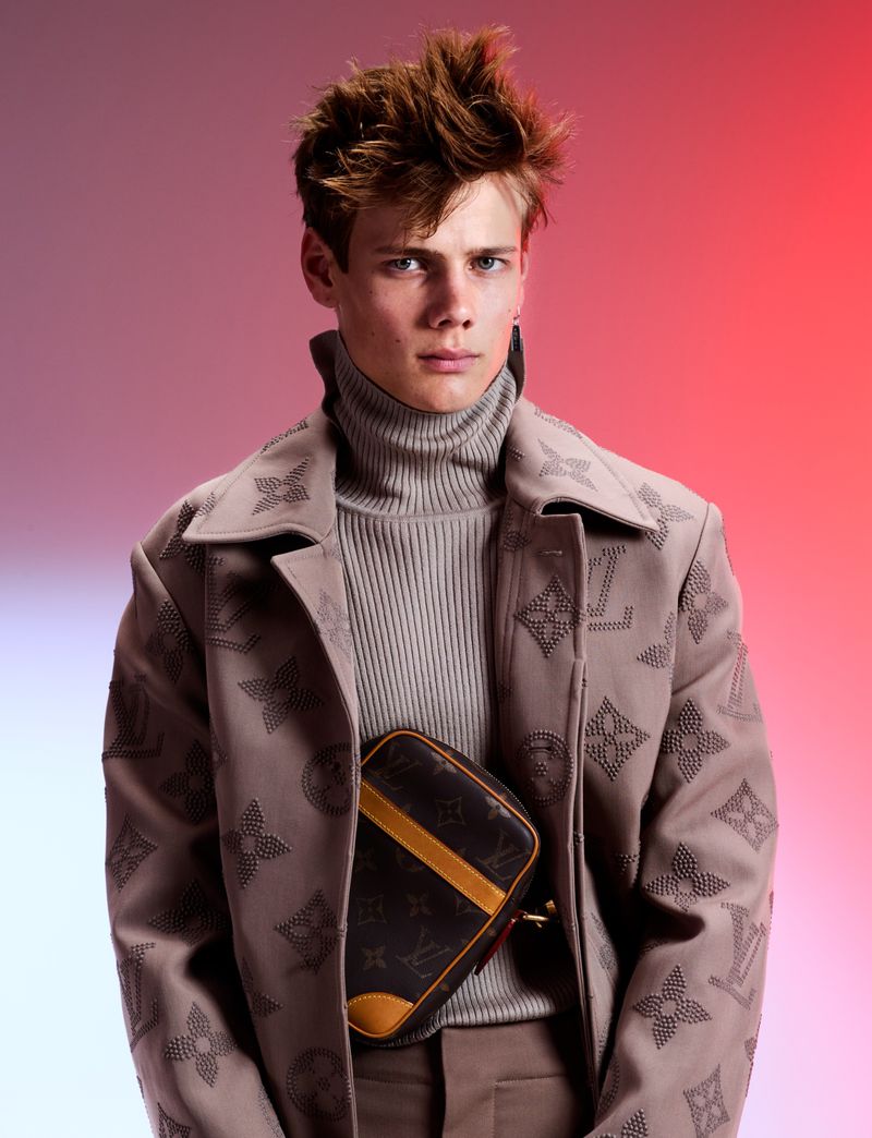 Stylish young adult man wearing a Louis Vuitton jacket and brown crossbody bag, posing against a pink and orange gradient backdrop.