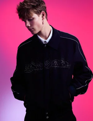 Young male model wearing a dark jacket with white stitching against a vibrant pink studio background.