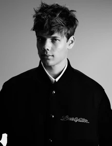 Portrait of a young male model with dark messy hair wearing a black bomber jacket against a plain grey background