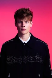 Portrait of a male model wearing a dark jacket over a white shirt against a pink gradient backdrop
