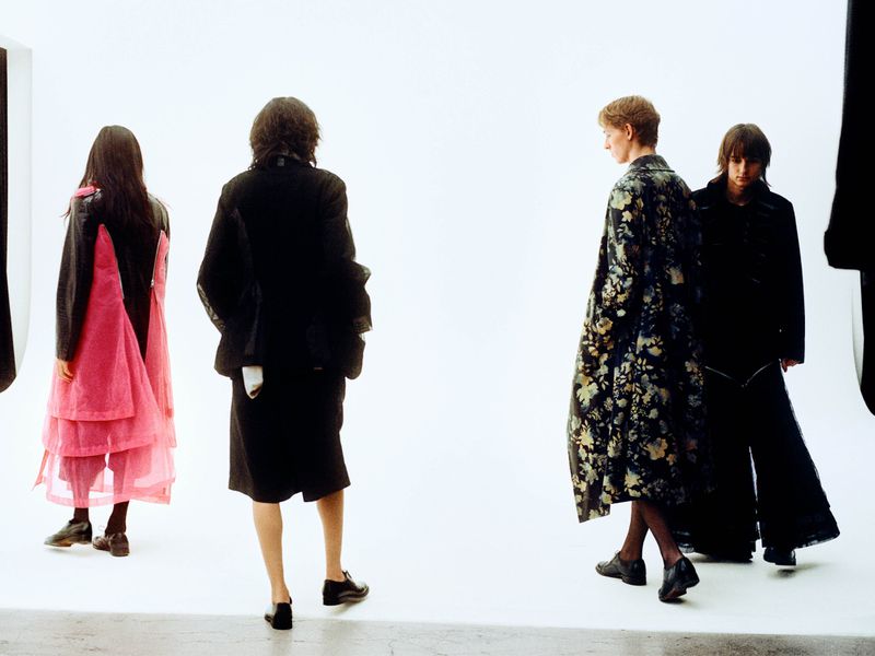 Four fashion models present womenswear in a studio setting. One model wears a pink tiered dress, while others wear black jackets and pants.