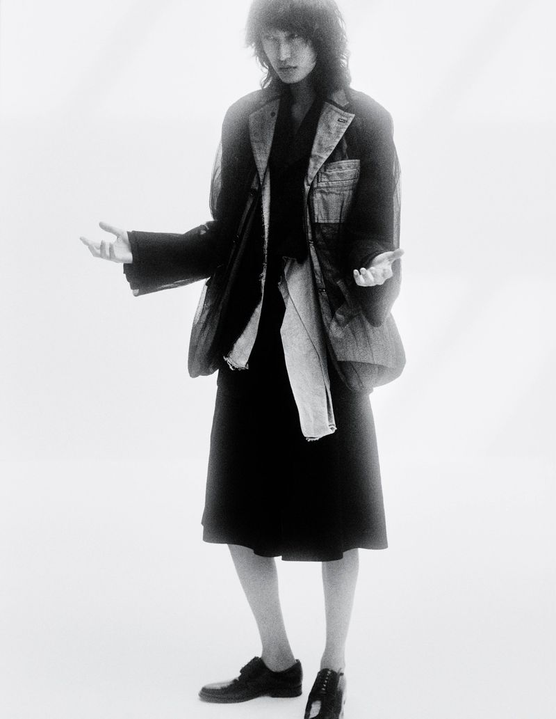 Fashion shot of a person wearing a blazer and skirt, standing against a white backdrop in a studio, creating a high-contrast black-and-white image.