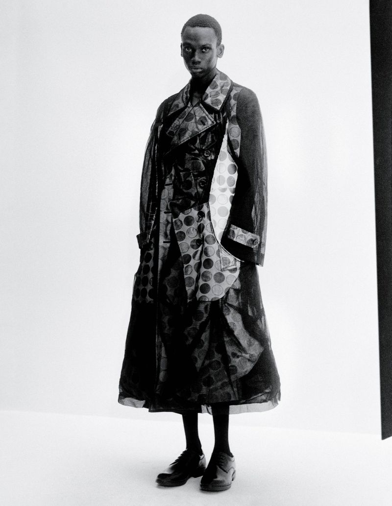 Monochrome portrait of a slender Black male fashion model in a long, patterned semi-sheer coat against a bright white backdrop in studio light.
