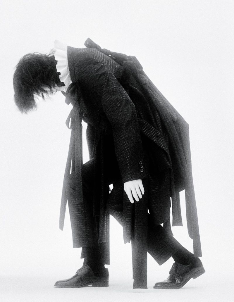 Monochrome fashion shot of a person leaning on a chair, wearing a suit, ruffle collar, and dress shoes in a bright, white indoor setting.