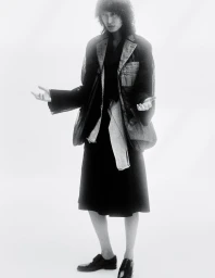 Model wearing dark layered jacket and skirt standing against a bright white studio backdrop with hands open