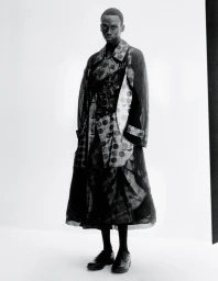 Black man wearing a patterned long coat posing against a seamless white studio background