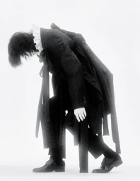 Model in layered black jacket and trousers leaning forward against a white background