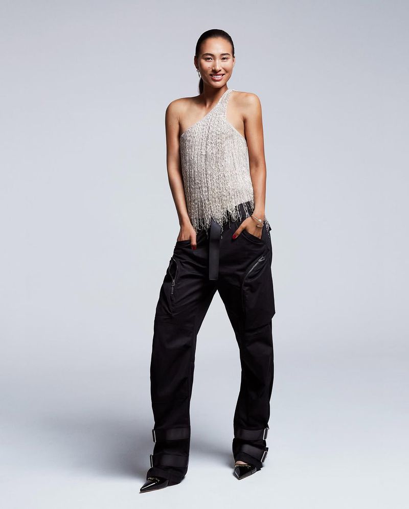 A woman in a white fringe top and black pants stands in front of a grey background. Fashion shot with a neutral background.