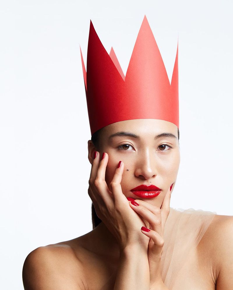 Glamour headshot of an Asian woman with a red paper crown, red lipstick, and matching manicure, resting her face in her hands on white.