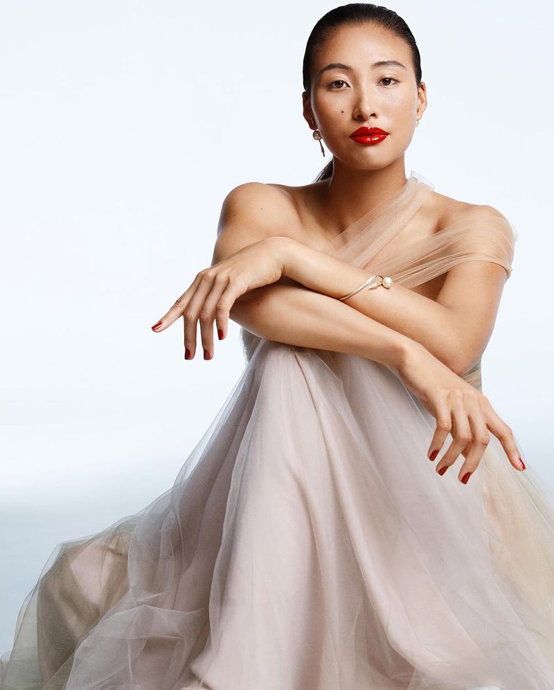 Elegant Asian woman with red lips and nail polish poses in a beige gown against a light backdrop, showcasing fashion and beauty.