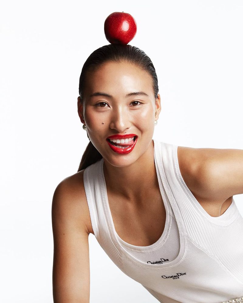 Zheng Qinwen balancing a red apple on her head while wearing a white ribbed tank top against a white background