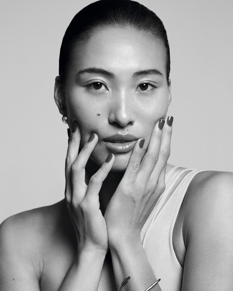 Close-up portrait of an Asian adult woman with her hands framing her face in a beauty shot against a neutral, monochrome background.
