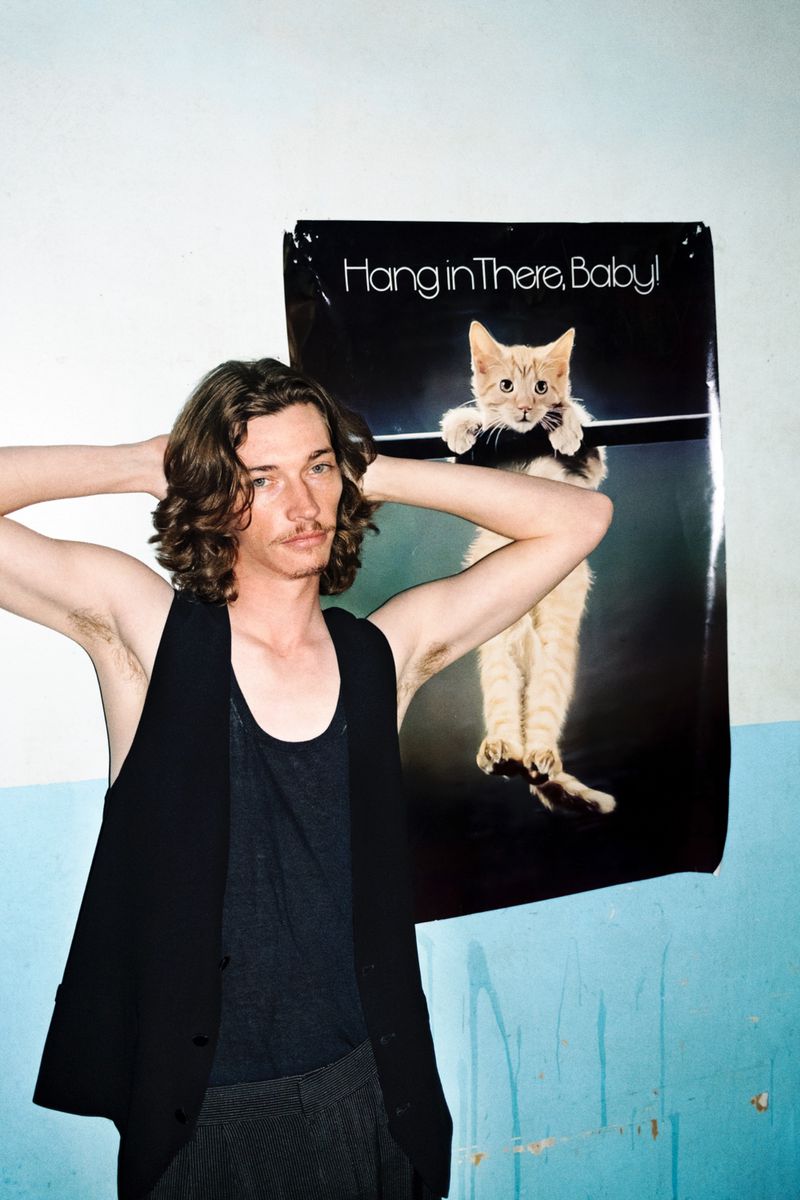 A man with long brown hair poses next to a vintage poster of a cat clinging to a pole with the phrase 'Hang In There, Baby!'