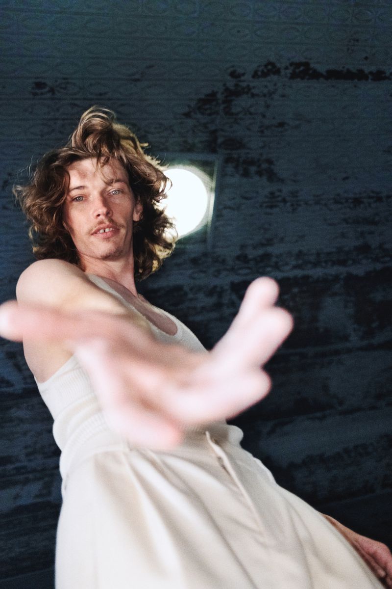 Low-angle shot of a man with long brown hair reaching out, wearing a white tank top and pants, with a ceiling in the background.