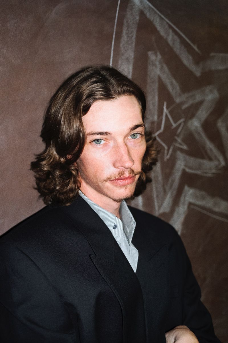 Close-up portrait of an adult male with long brown hair, mustache, wearing a black suit with a light-grey button up shirt, against a brown background.