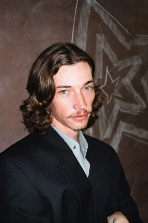 Close-up portrait of an adult male with long brown hair, mustache, wearing a black suit with a light-grey button up shirt, against a brown background.