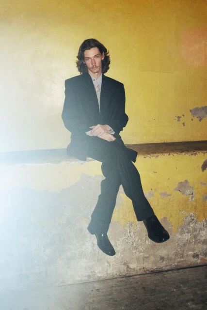 Adult male with long brown hair wearing a suit sitting against a yellow wall looking at the camera in a medium shot indoors.