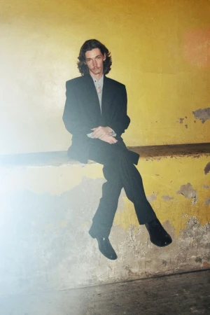 Adult male with long brown hair wearing a suit sitting against a yellow wall looking at the camera in a medium shot indoors.