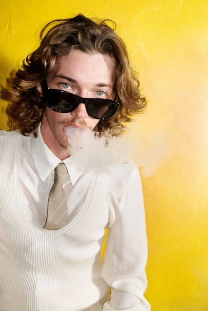 Adult male in sunglasses exhales vapor against a yellow backdrop, styled in a white shirt, tie, and vest for a modern, edgy aesthetic.