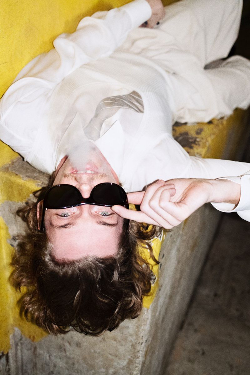 Upside-down portrait of an adult male reclining on a yellow surface, smoking with sunglasses and casual off-white clothing.
