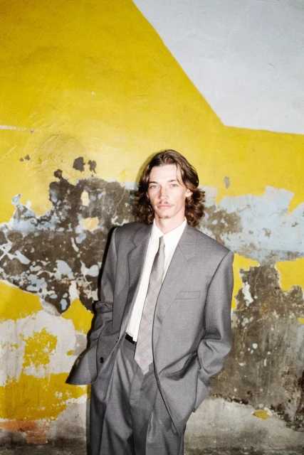 Man in gray suit stands against weathered yellow and blue painted wall, hands in pockets, looking at the camera in portrait orientation.