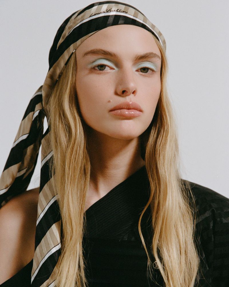 Close-up portrait of a woman with light blonde hair, light green eye shadow, and a striped headscarf against a white background.