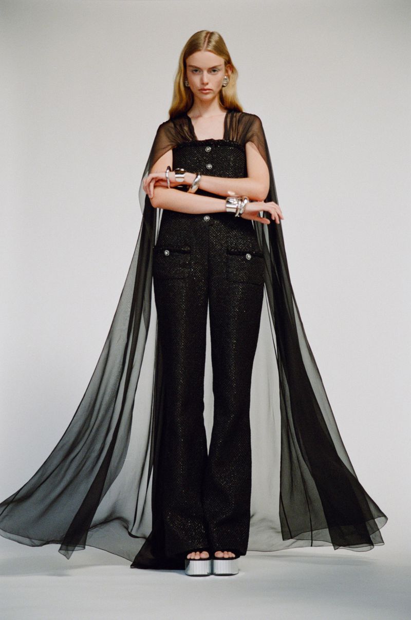 An adult female fashion model poses in a shimmering black jumpsuit and sheer black cape in a brightly lit photo studio.
