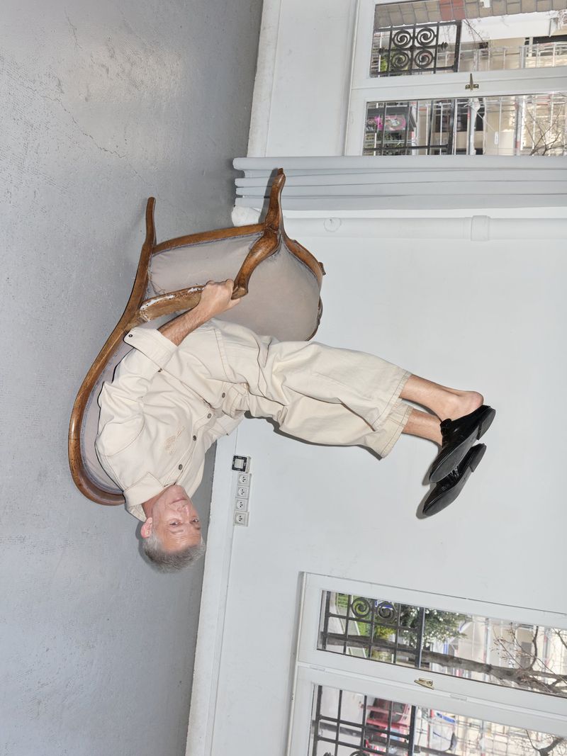 Mature adult balancing upside down on a chair indoors, wearing a beige outfit and black shoes, in front of a window.