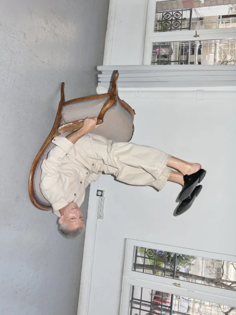 Mature adult balancing upside down on a chair indoors, wearing a beige outfit and black shoes, in front of a window.