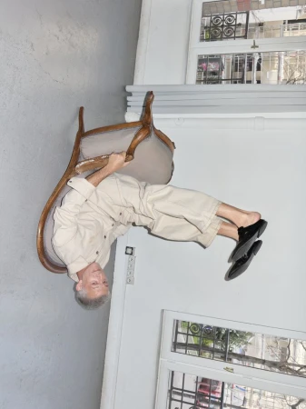 Mature adult balancing upside down on a chair indoors, wearing a beige outfit and black shoes, in front of a window.