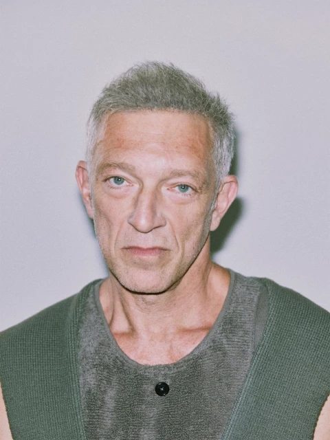 Portrait of an adult male with grey hair wearing a sleeveless top with a button, set against a plain white background.