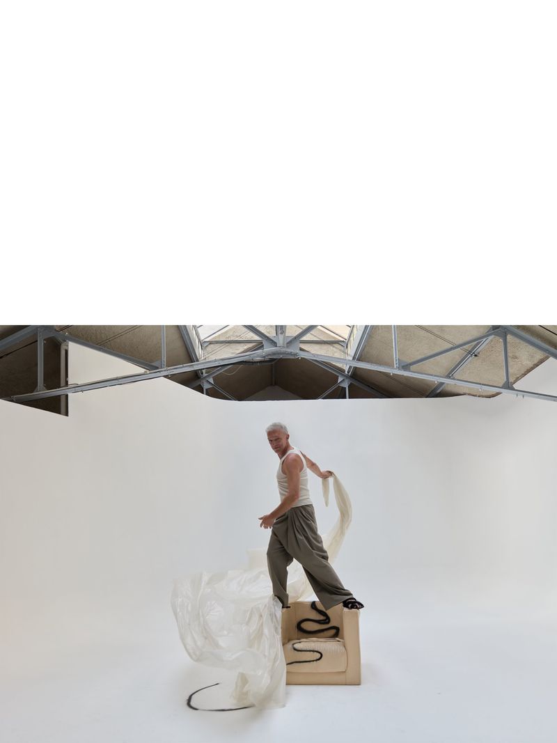 Adult male in a photo studio stands on a wooden cube with a handbag and flowing white cloth, posing under natural skylight.