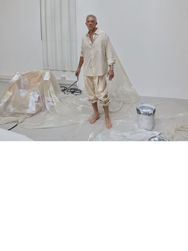 Full shot of an adult man standing in a white room with a bucket and paint tarp, posing for the camera in relaxed posture.