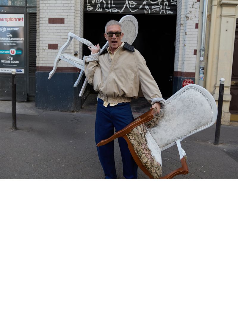 Adult male carries two white chairs outside building with ad signage; he wears a light jacket, dark pants, and glasses.