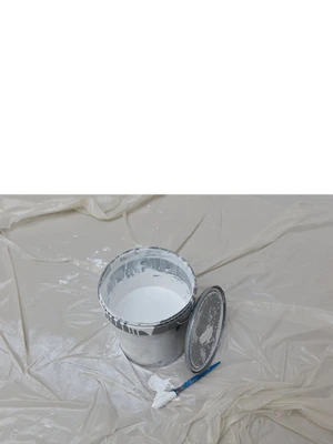 An open bucket of white paint with its lid beside a paintbrush on plastic sheeting