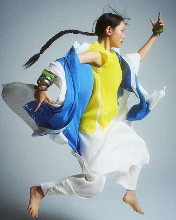 Asian woman in mid-air in a yellow top, white pants, and a blue and white shawl, accessorized with green bracelets in a studio.