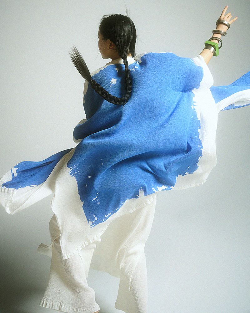 Back view of an Asian adult female wearing a blue-and-white abstract-patterned robe and pants, with her arms raised, accessorized with metal bracelets.